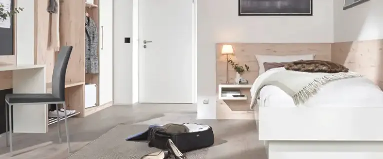 Helles Schlafzimmer mit weissem Bett vor Holzrückwand und Nische mit Lampe, links offener Garderobenschrank und Stuhl, vorn geöffneter Koffer auf Teppichläufer.